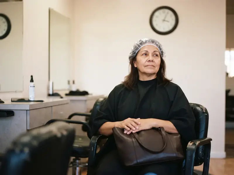 A woman waits alone in a salon chair with her color processing under a plastic cap, looking up at the wall clock