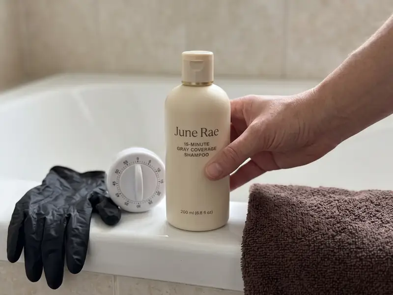 The June Rae shampoo bottle on the edge of a bathtub beside a white kitchen timer, a dark towel and black gloves